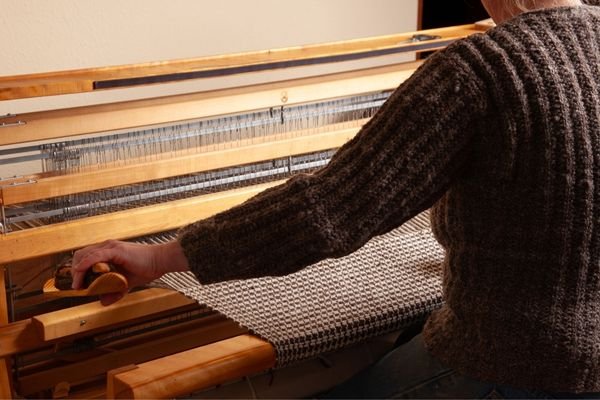 a woman in a brown sweater weaving at a floor loom