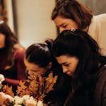 Group of women arranging flowers with autumnal leaves indoors, showing creativity and concentration.